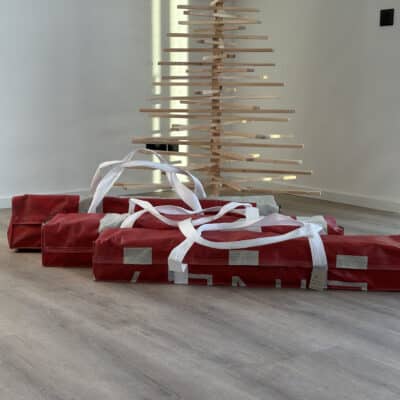 Drei rote Upcycling-Taschen von Fyksin für die Aufbewahrung der Franky Tree Holzweihnachtsbäume, hergestellt aus alten Werbebannern, vor einem aufgebauten Franky Tree.